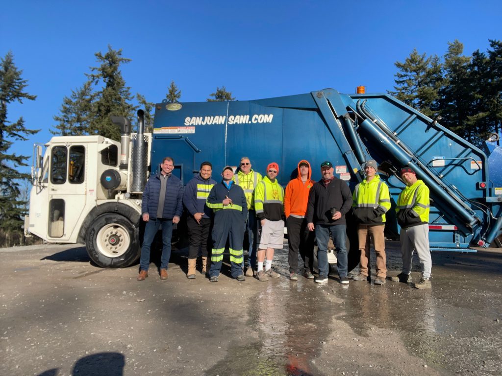 A group of employees poses in front of a San Juan Sanitation garbage truck