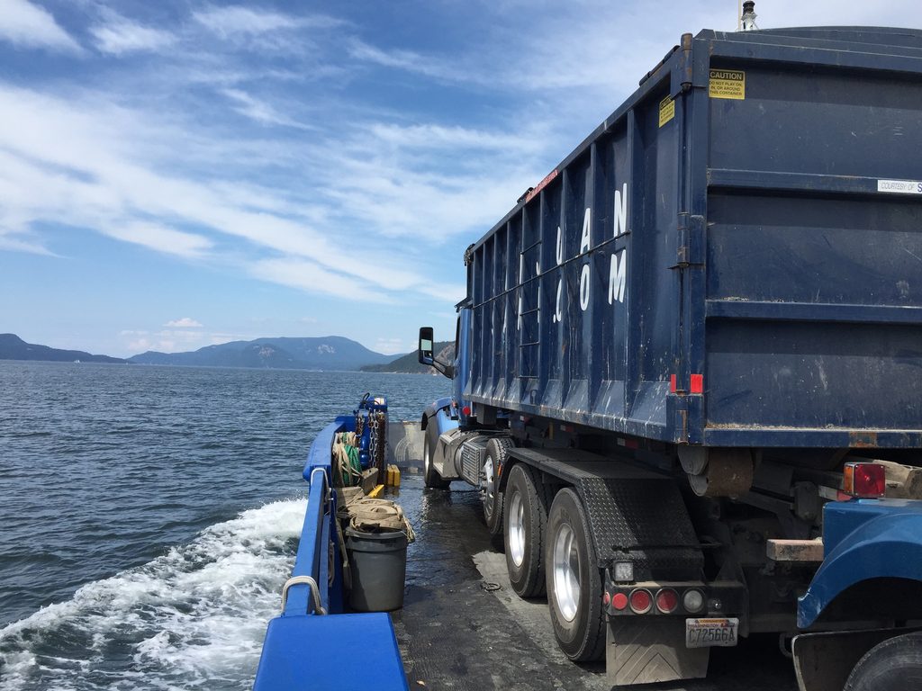 a blue garbage truck sits on a barge in the puget sound