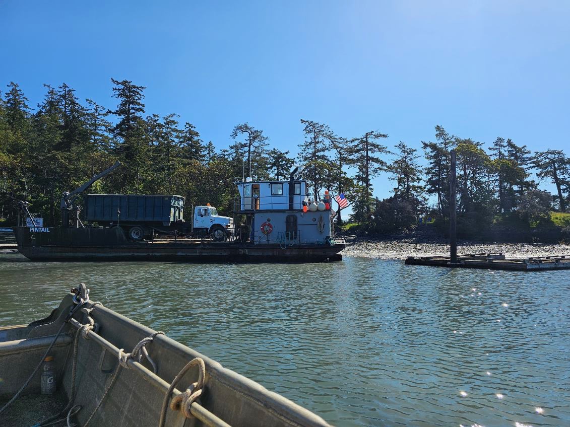A garbage truck sits on a barge on a sunny day in the San Juan islands